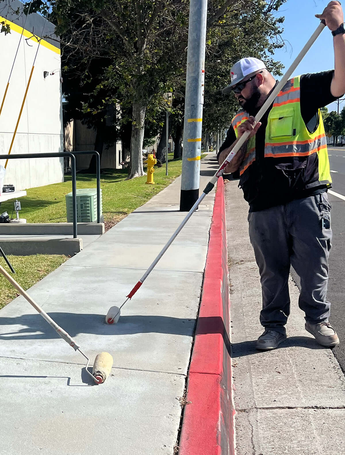 Like Nu Commercial Services worker painting curbside with a long-handled roller brush in a safety vest and helmet.