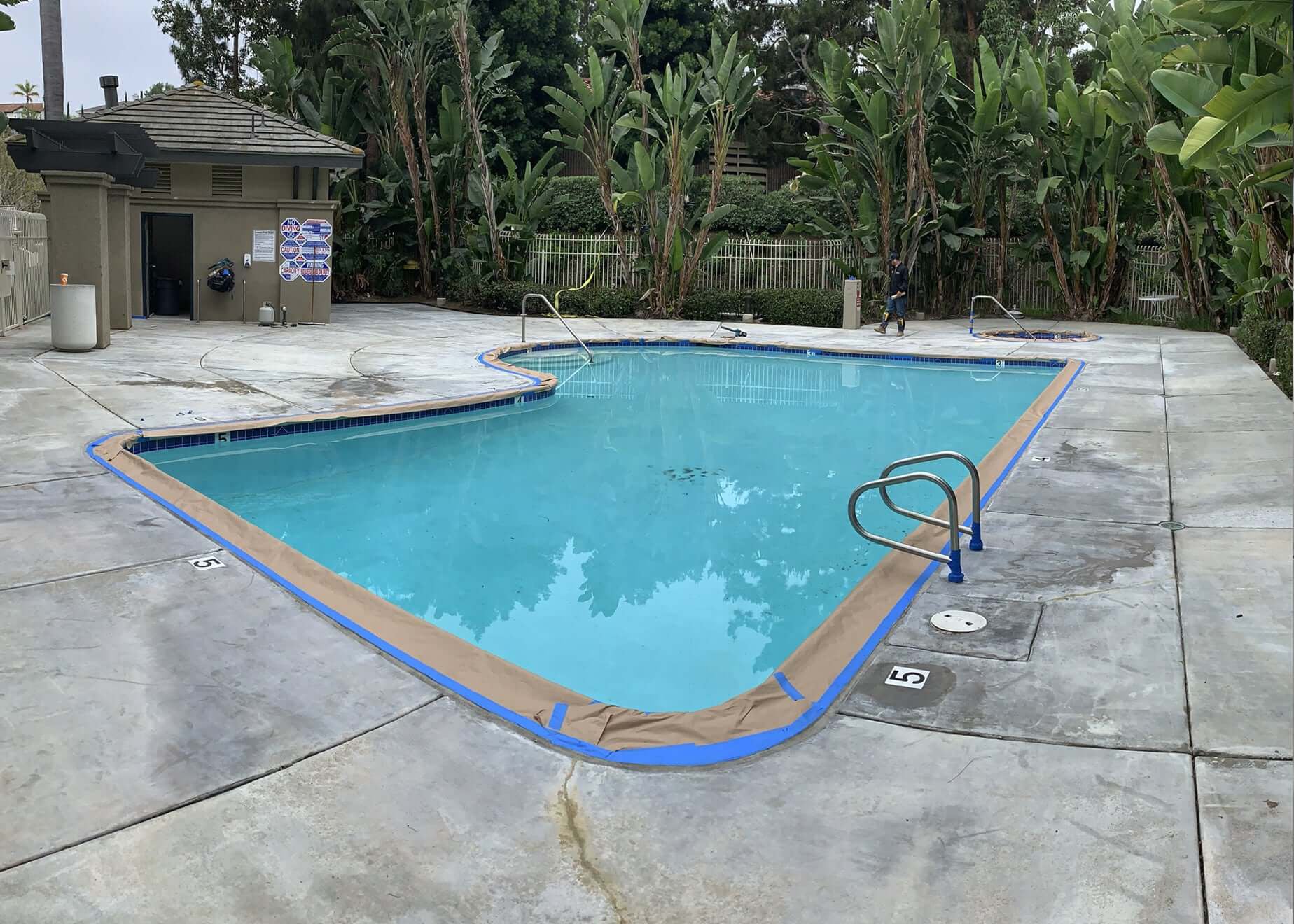 Swimming pool area undergoing concrete restoration with tape marking the edges, surrounded by lush greenery.